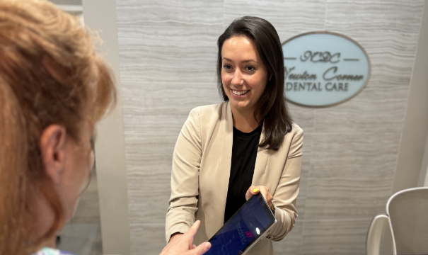 A dental receptionist in a beige blazer smiles while handing a tablet to a patient. The office sign in the background reads 'NCDC - Newton Corner Dental Care'.